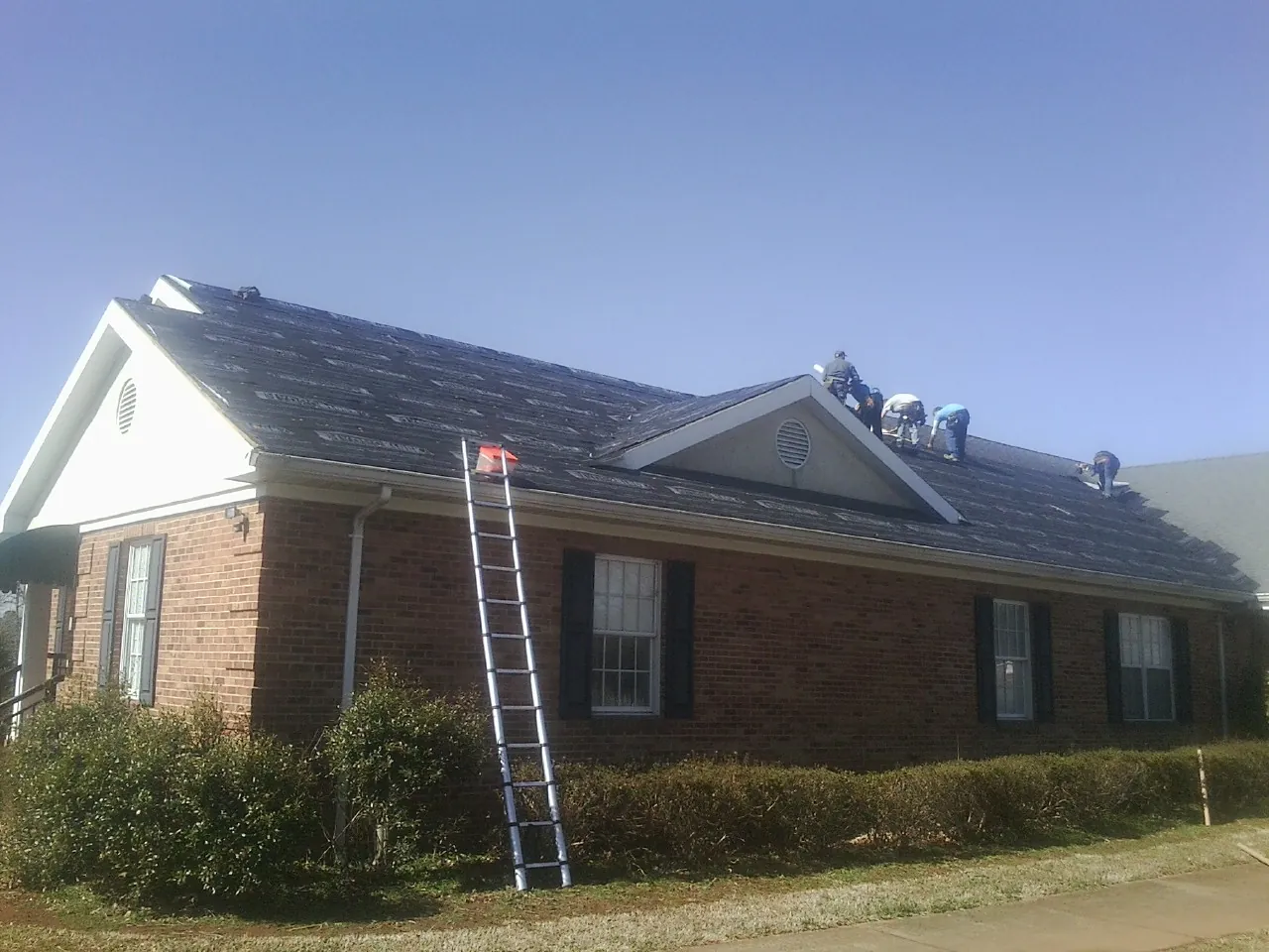 Crew tearing shingles off a brick ranch roof with a ladder set — Emergency Roof Repair work by our crew in Hope Mills