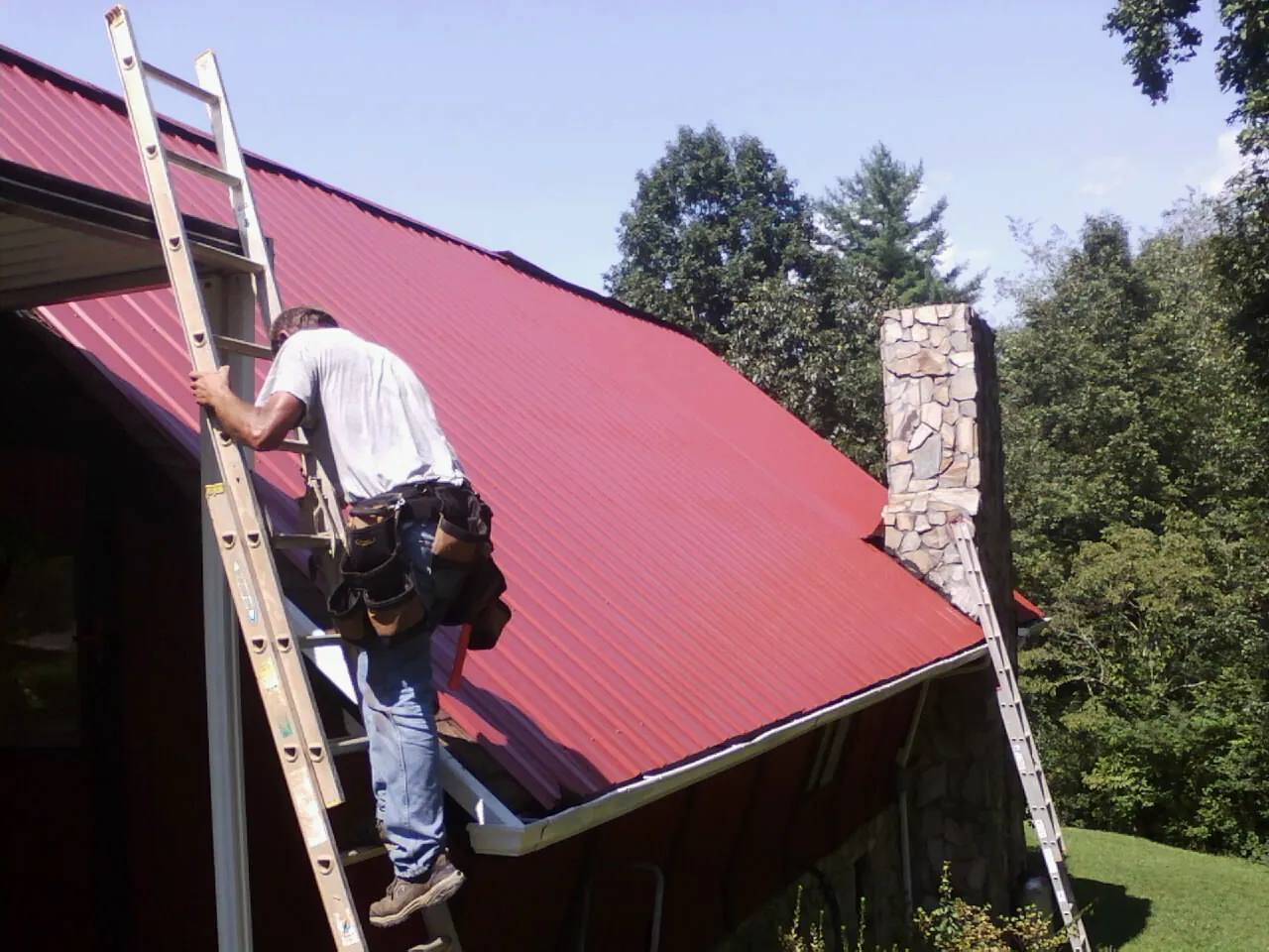 Roofer on a ladder beside a red metal roof and stone chimney — Soffit & Fascia Repair by Roofing Brothers in Hope Mills